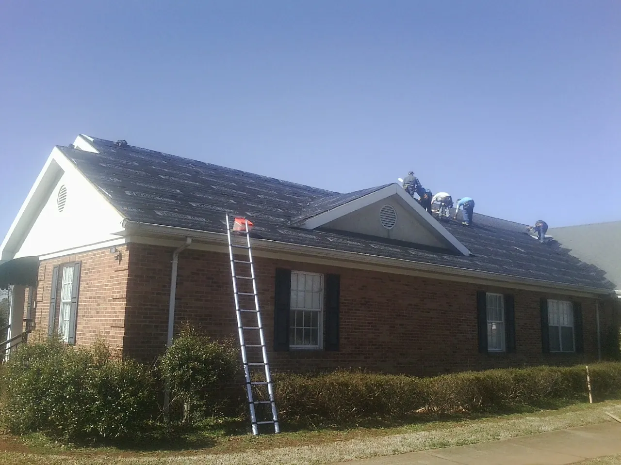 Crew tearing shingles off a brick ranch roof with a ladder set — Emergency Roof Repair work by our crew in Cottage Grove
